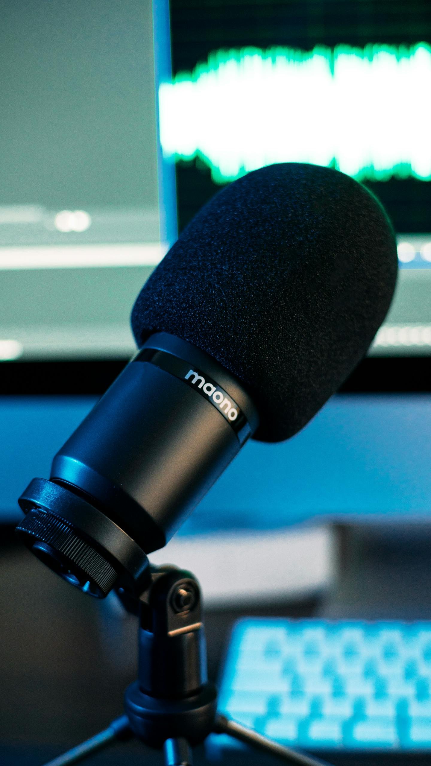 Close-up of a black microphone in a music studio with sound waves on a monitor in the background.